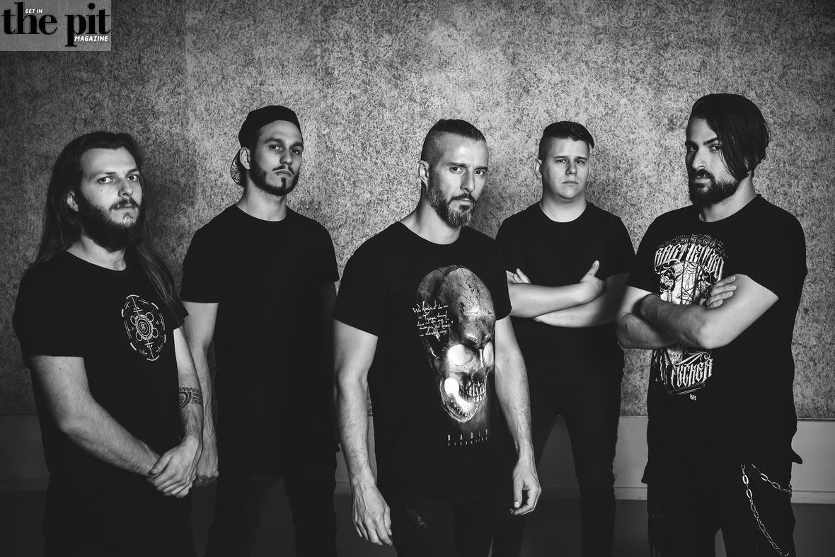 Five disconnected men standing against a gray wall, dressed in black t-shirts, looking seriously at the camera.
