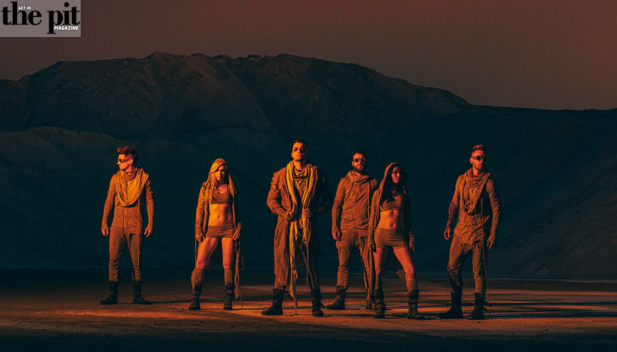 Six people in rugged outfits standing in a desert under a sunset, with the logo "the pit magazine" on the top left.