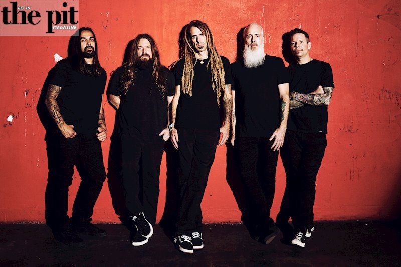 Five members of the band Lamb of God pose against a red wall, dressed in black with distinct hairstyles and tattoos.