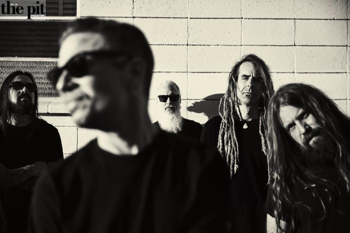 Four members of the rock band Lamb of God, with one in focus at the forefront, against a brick wall background, providing a stark black and white contrast.