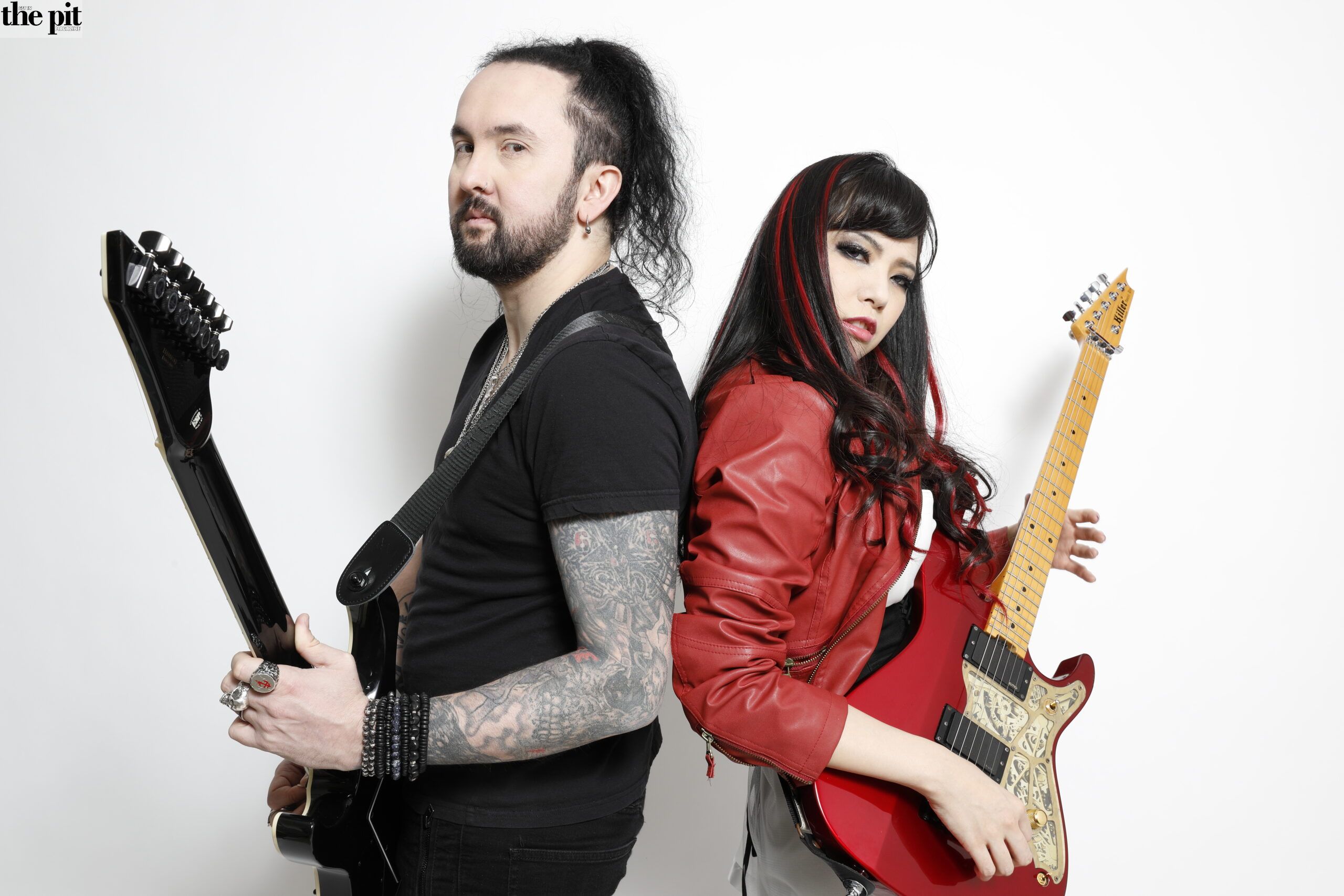Two musicians posing with electric guitars from Amahiru, one man with a black shirt and tattoos and one woman in a red jacket.