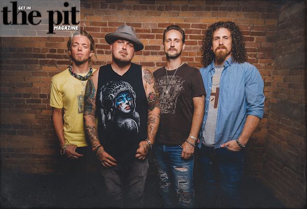 Four men standing against a brick wall, each styled in casual, Black Stone Cherry rocker attire, posing confidently for the camera.