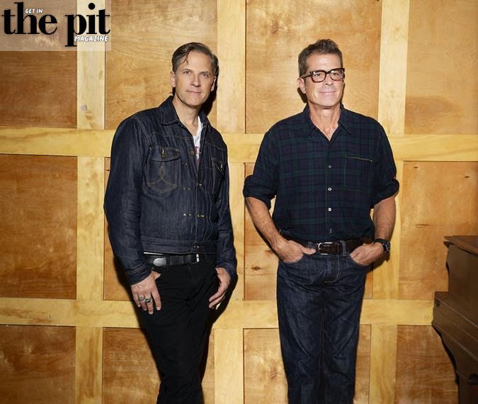 Two mature men in denim and plaid shirts standing against a wooden backdrop with a Calexico magazine logo "in the pit" at the top left corner.