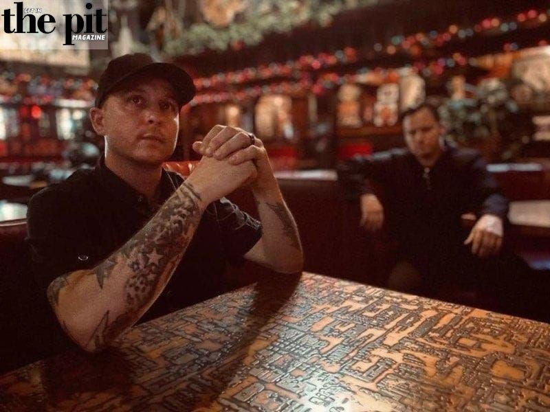 Two men sitting at a bar, one in the foreground with tattoos on his arms and a Royalty Kult cap, and the other in the background, in a dimly lit room with eclectic decor
