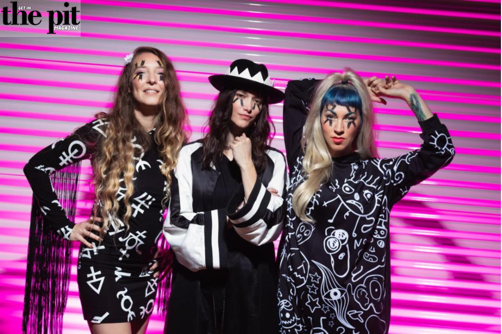Three women in patterned outfits pose confidently on stage with a vibrant pink striped background, representing The Dead Deads.