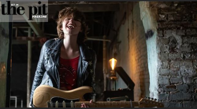 A joyful young musician with a guitar, wearing a leather jacket, laughs in a rustic, brick-walled room at Jax Hollow.