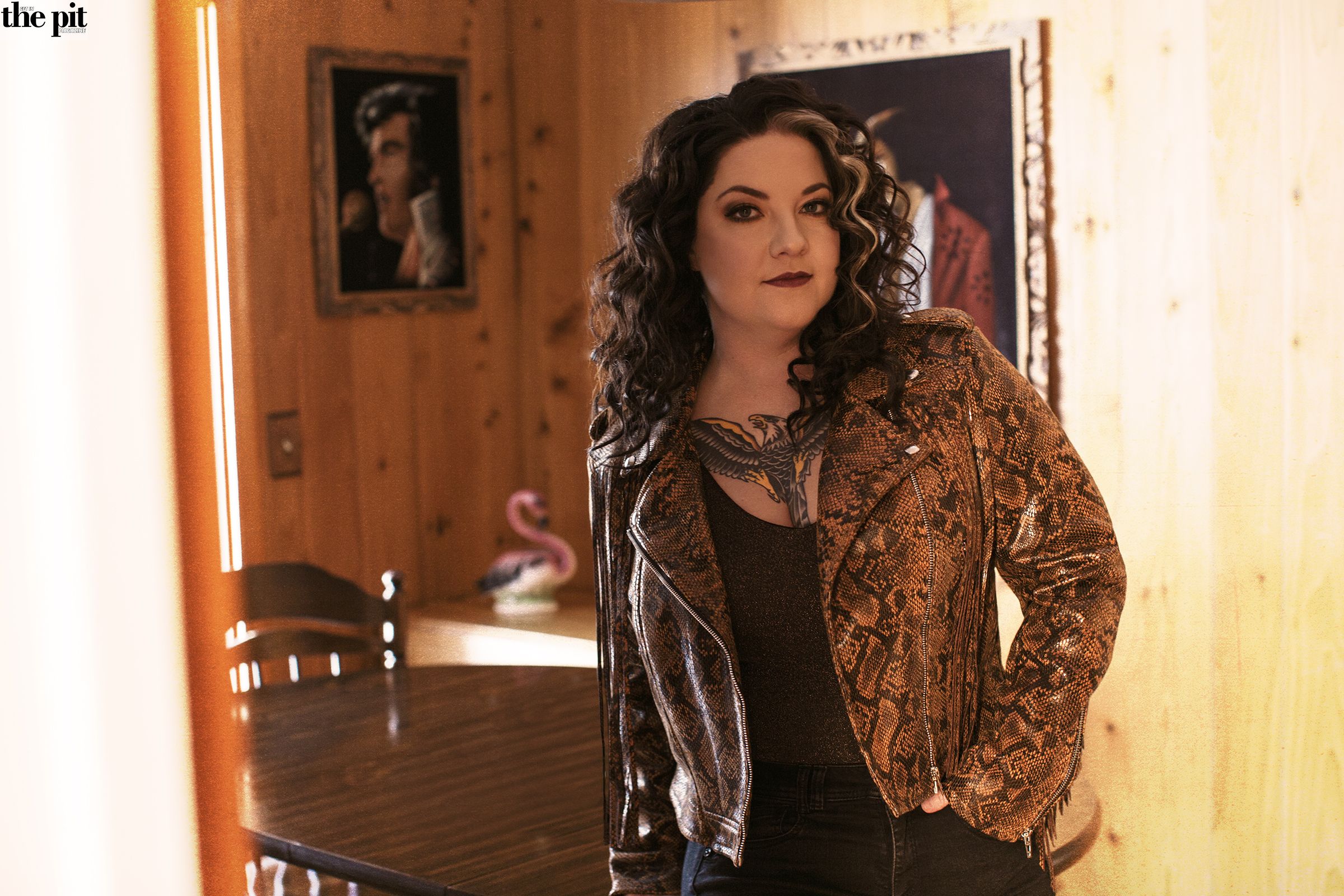 Ashley McBryde stands confidently in a room with wood-paneled walls, wearing a patterned jacket and jeans.