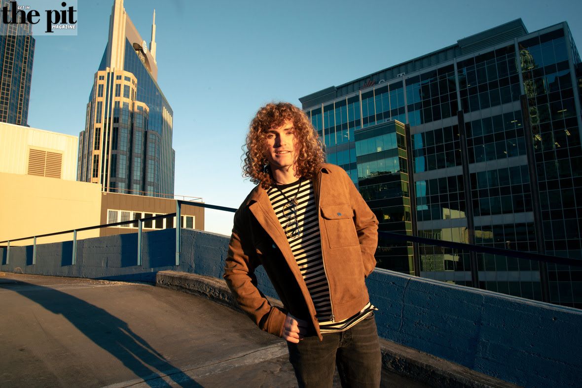 A man with curly hair standing on a rooftop, wearing a brown jacket, with modern buildings and a pointed skyscraper in the background during the filming of "Eddie and The Getway.