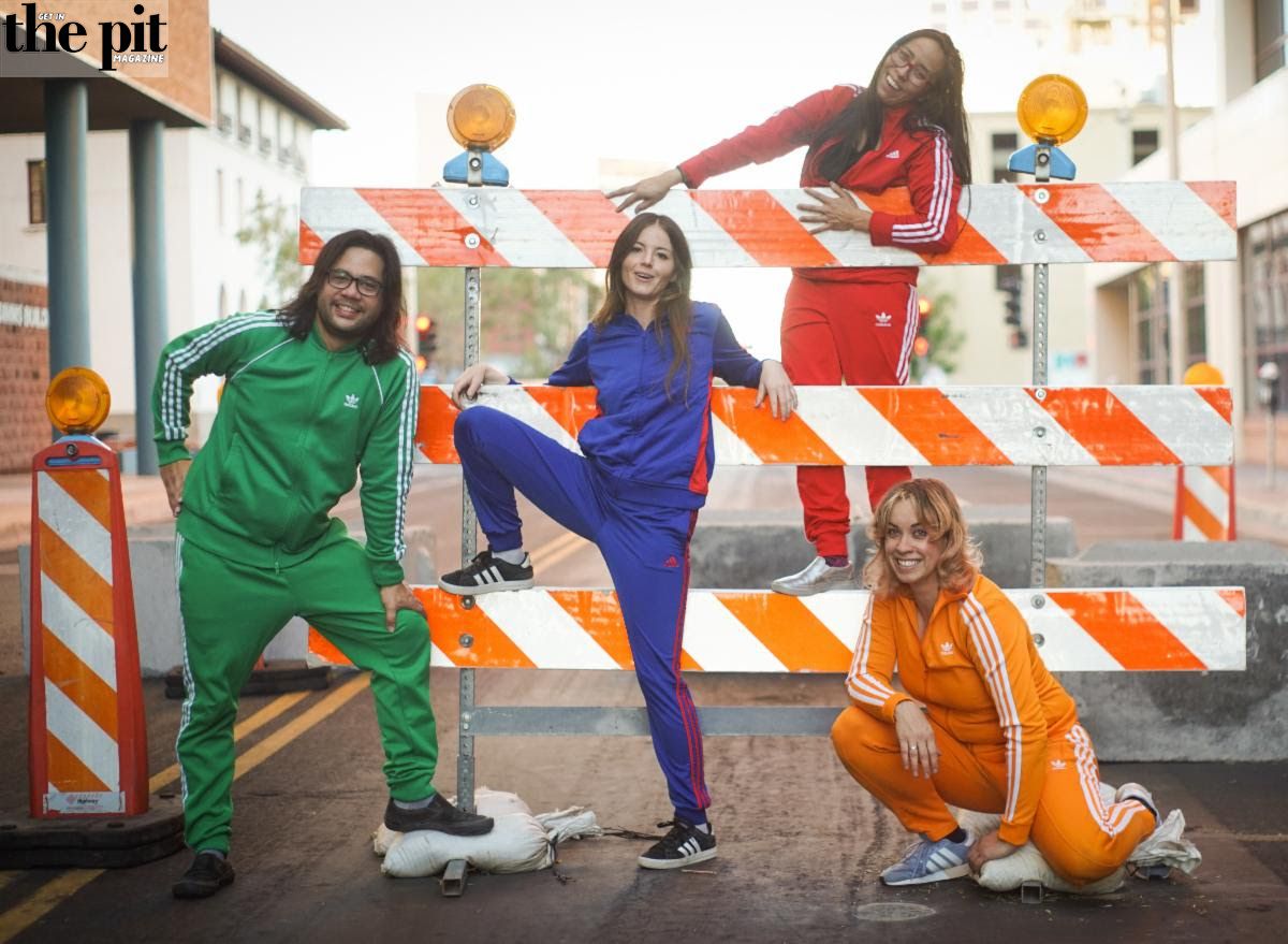 Four people in colorful tracksuits pose playfully on and around an orange roadblock barrier in an urban setting, embodying the vibrant energy of Prism B!tch.