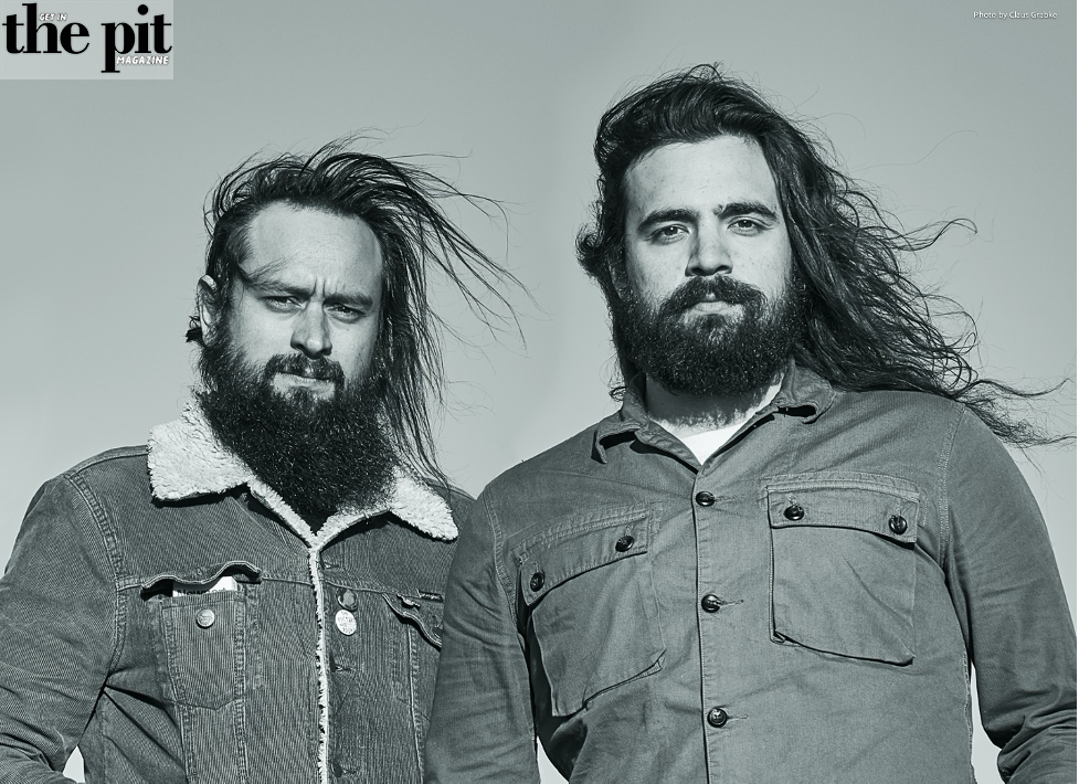 Two men with long hair and beards, dressed in denim and buttoned shirts, posing for a black and white portrait.