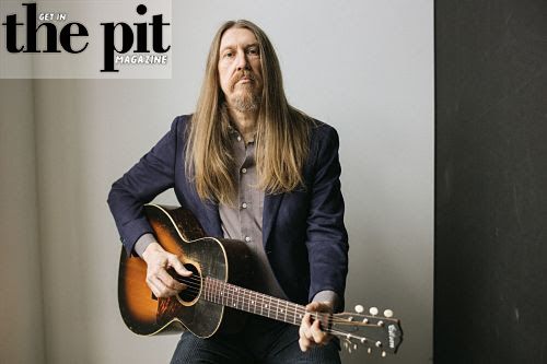 Oliver Wood, with long hair and beard, wearing a navy blazer, playing an acoustic guitar, seated against a gray background.