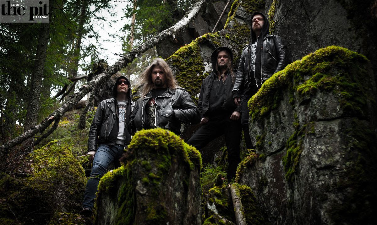 Four men in leather jackets, including one hooded menace, standing among mossy rocks and trees, exuding a rugged outdoor style.