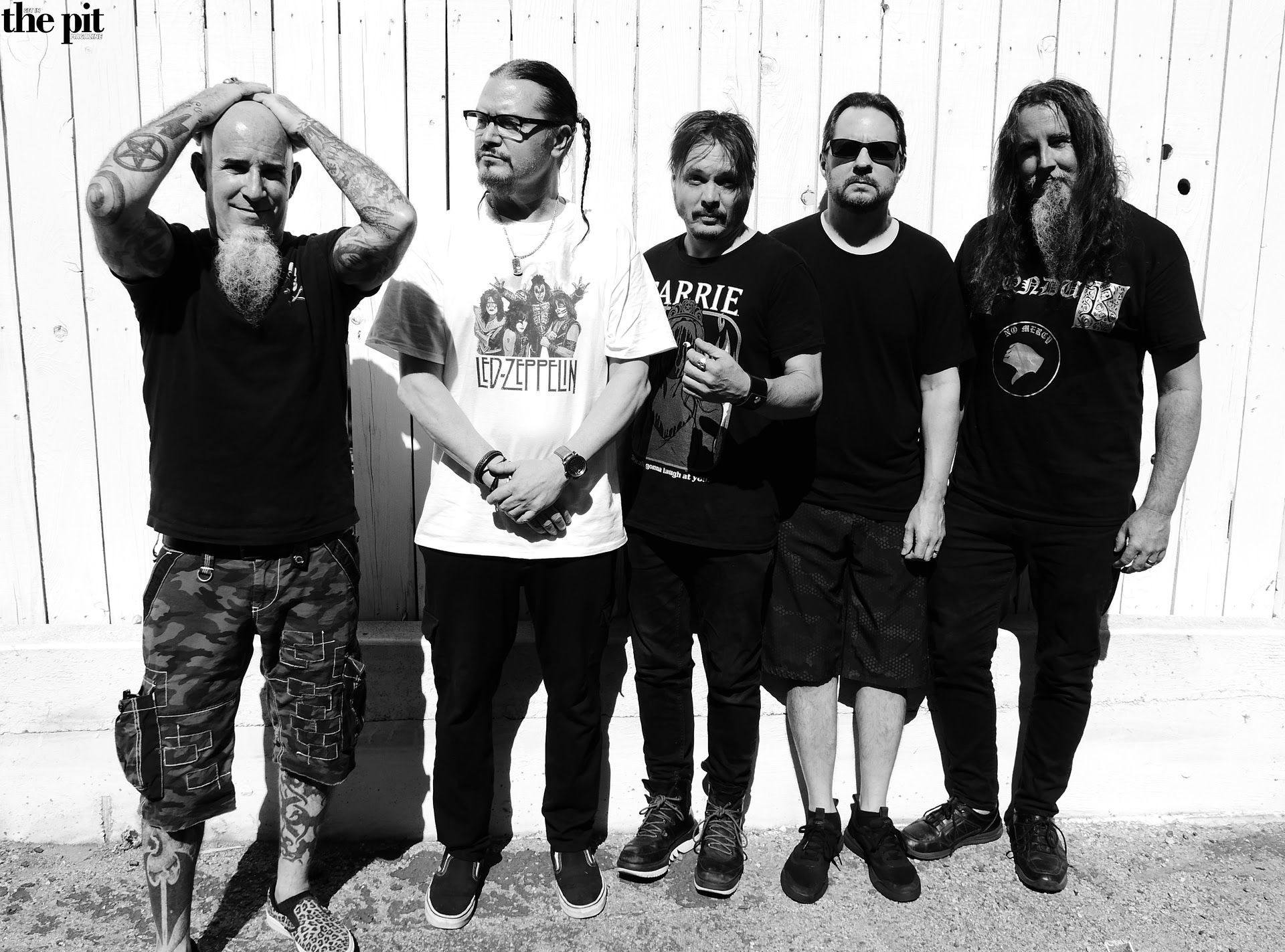 Five men standing in a line against a wooden wall, dressed in casual black clothing with visible Mr. Bungle t-shirts, posing for the camera.
