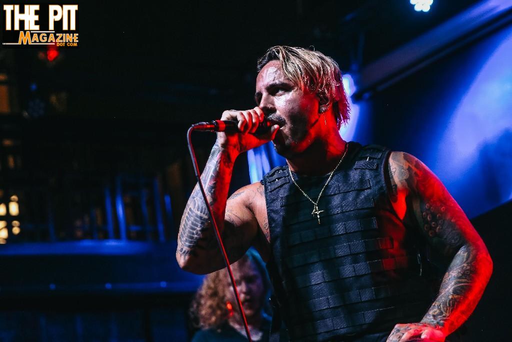 A tattooed male singer from Ovtlier performing onstage with a microphone, wearing a black vest, and the logo of "the pit magazine" in the corner.