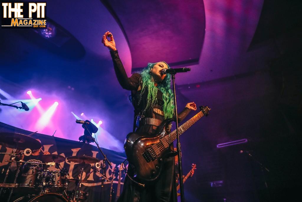 Female rock musician with green hair playing a guitar and singing passionately on stage under colorful stage lights with Stitched Up Heart.