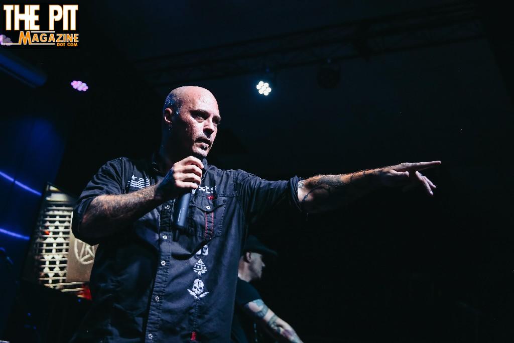A bald man with tattoos, wearing a dark vest, performs on stage as part of A Killer's Confession, speaking into a microphone and pointing to the side. The background features dim lighting and blurred