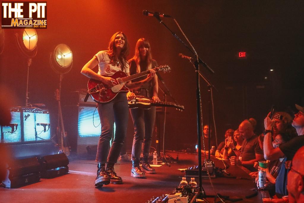 Two female musicians, known as Larkin Poe, performing on stage, one playing a guitar and the other on a keyboard, with an audience in the foreground.