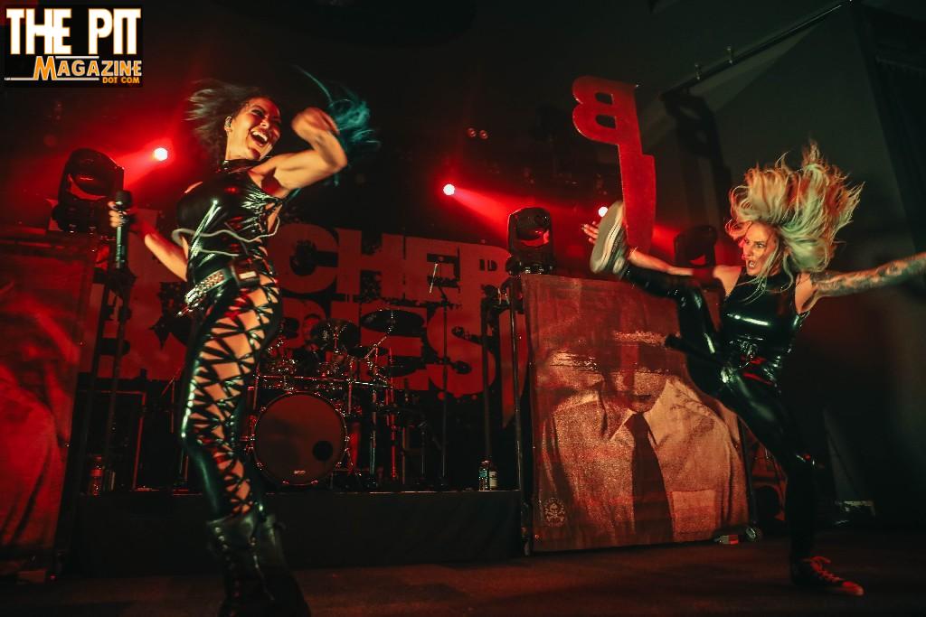 Two energetic performers from Butcher Babies, with blue and blonde hair respectively, jumping energetically on stage during a live concert, under red lighting.