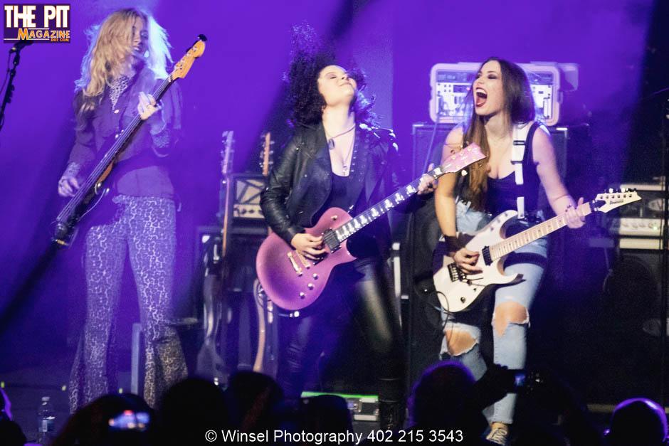 Three female musicians on stage, one singing and two playing plush guitars, with vibrant stage lighting in the background.
