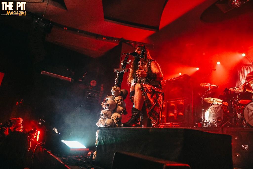 A musician in elaborate tribal costume performs on stage with teddy bears attached to his microphone stand, under red stage lighting, evoking the intense spirit of Black Label Society.