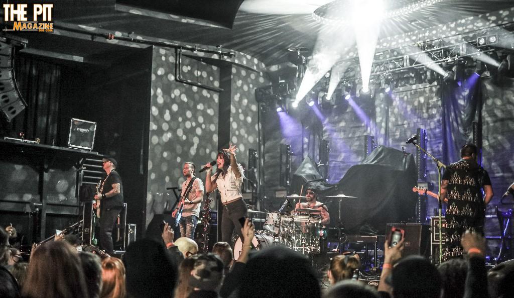 A band performs live on stage at a crowded concert venue with energetic audience and stage lights.
