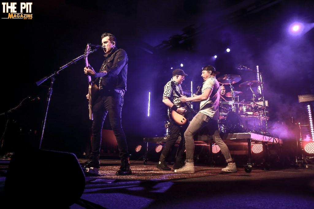 Band Theory of a Deadman performing on stage with a singer, two guitarists, and a drummer, illuminated by dramatic purple and blue stage lights.