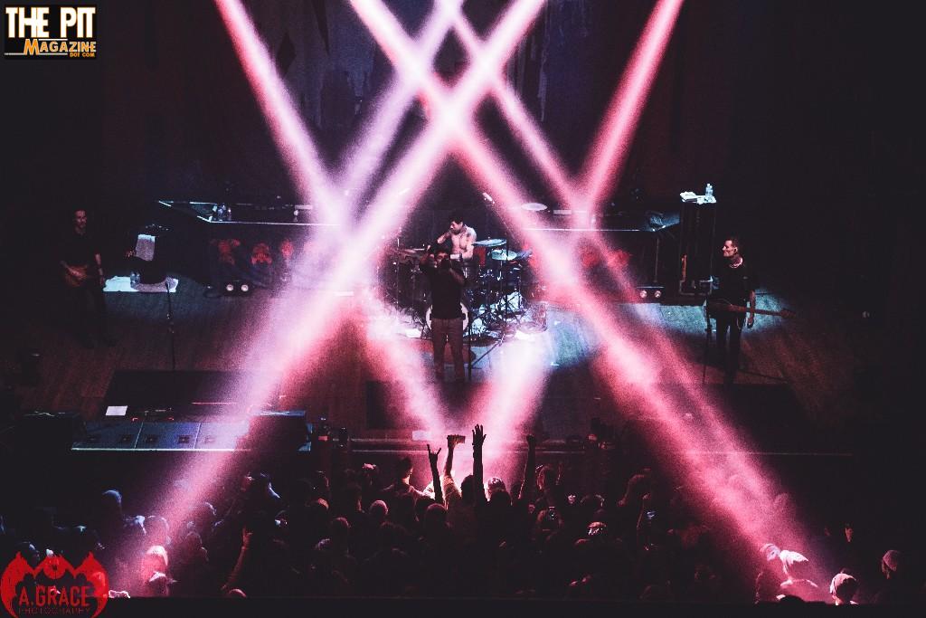 Crown the Empire performs on stage under striking pink and white lights, with an enthusiastic crowd visible in the foreground.