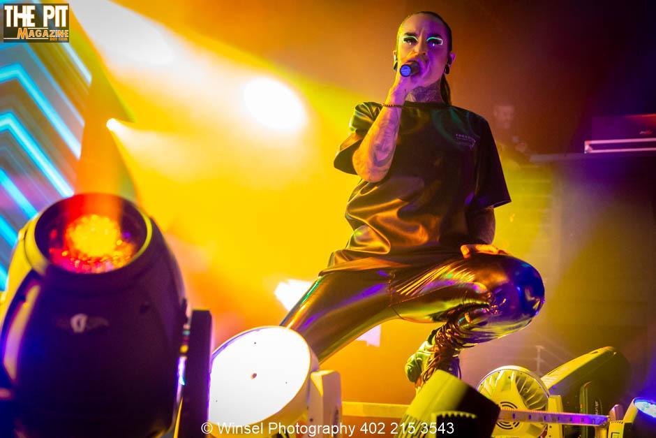 Bald performer with facial tattoos from Jinjer sings into a microphone on stage, illuminated by vibrant yellow stage lights.