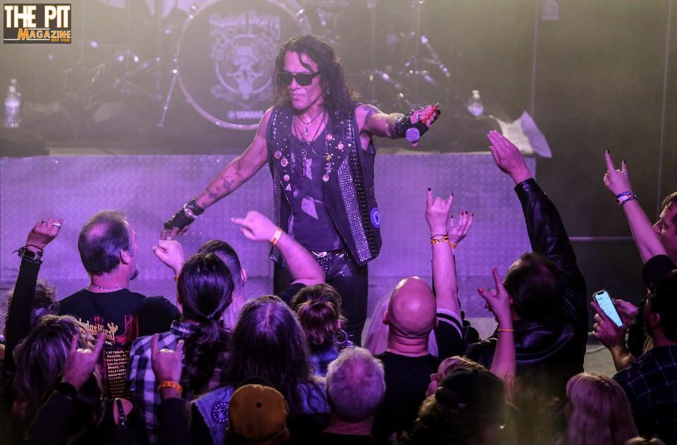 Stephen Pearcy performing on stage, wearing sunglasses and leather, with enthusiastic crowd raising hands and recording with phones.