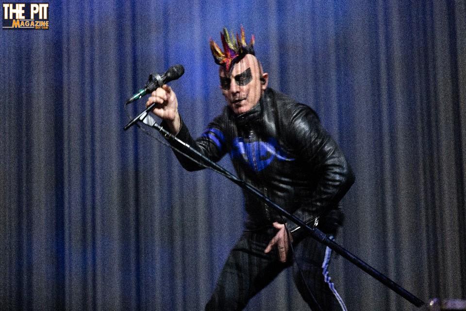 A punk rock singer performing on stage with a spiked mohawk and leather jacket, holding a microphone. The background has a blue curtain and a Tool magazine logo.