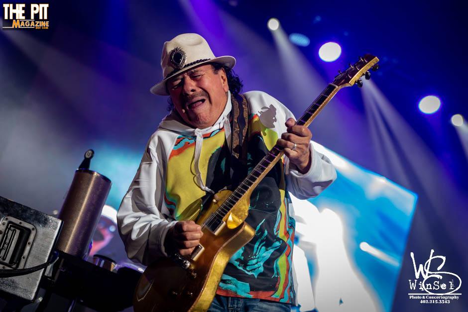 Santana, a guitar player in a white hat and colorful shirt, performs enthusiastically on stage under blue lights.