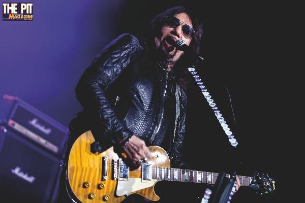 Rock musician Ace Frehley in a leather jacket, performing on stage, singing into a microphone and playing a yellow electric guitar.