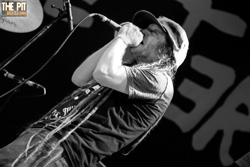 A black and white photo of a male singer from Circle Jerks in a hat, performing passionately into a microphone onstage.