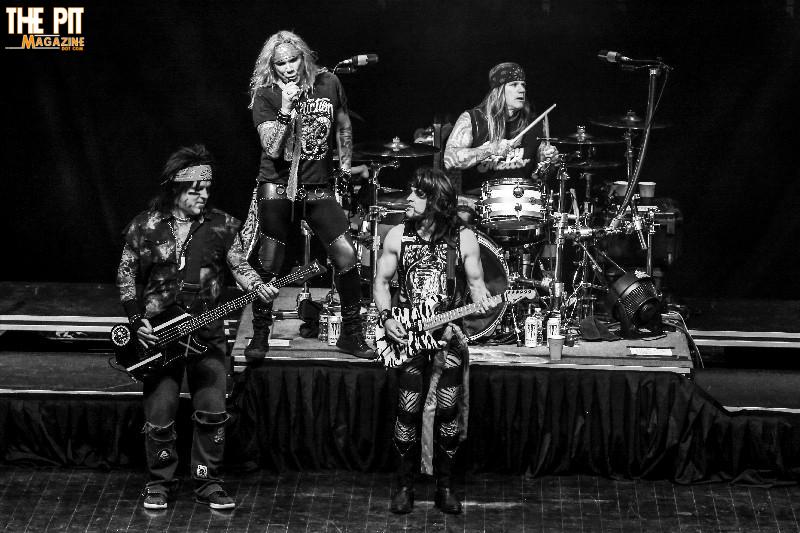 Black and white photo of Steel Panther performing on stage, featuring four band members playing guitar, bass, and drums.
