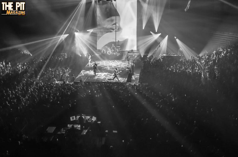 Black and white photo of a lively Shinedown concert, showcasing the band performing on stage with dramatic lighting while the audience raises their hands in excitement.