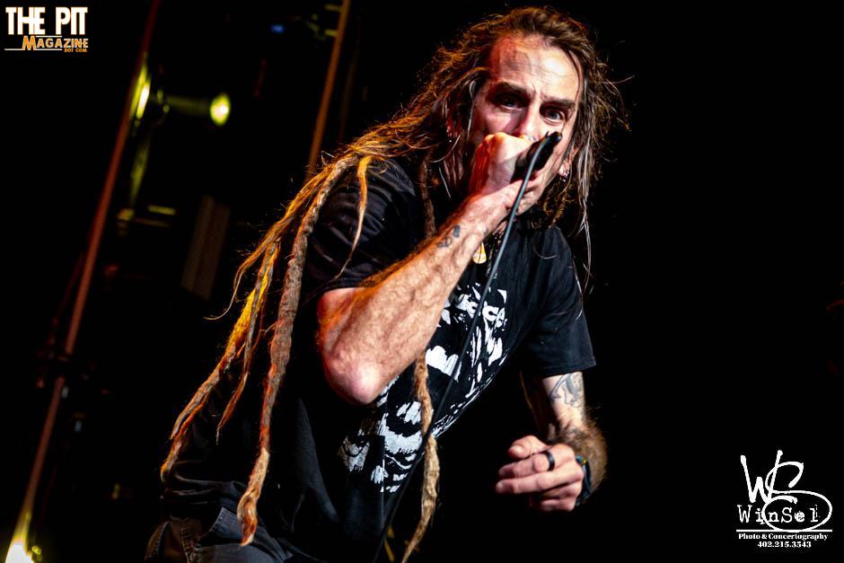 A male rock vocalist from Lamb of God with dreadlocks and a black shirt performs energetically into a microphone on a concert stage, highlighted by stage lighting.