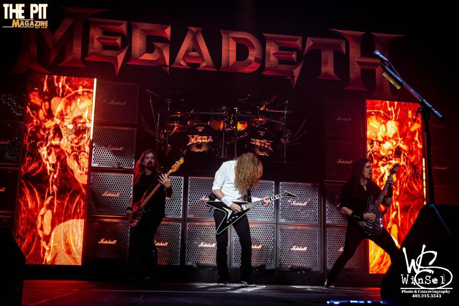 Megadeth performs on stage with fiery graphics in the background, featuring the band members playing guitars and drums.
