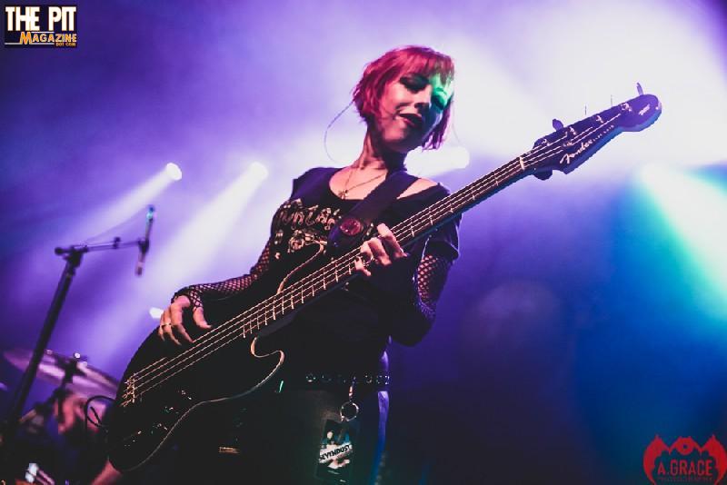 A female bass guitarist performing on stage, illuminated by blue stage lights, with a focused expression.