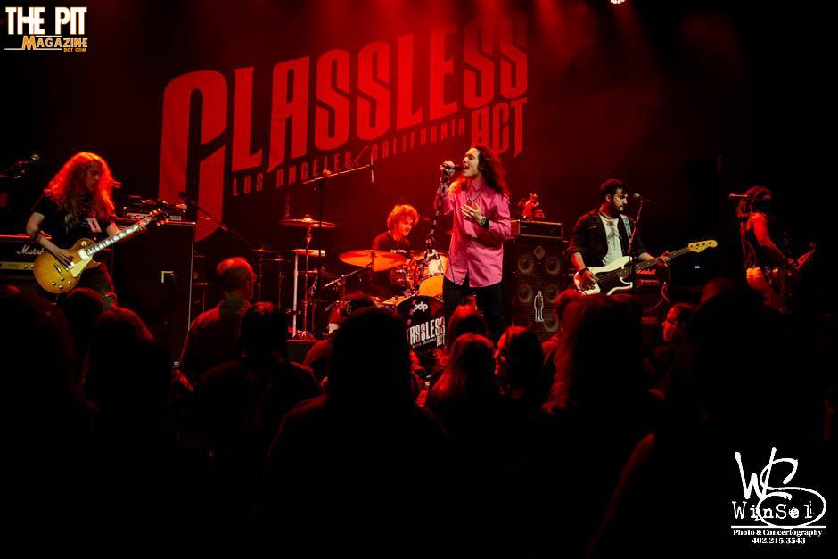Rock band performing on stage under red lighting with a backdrop reading "Classless Act" in the background.