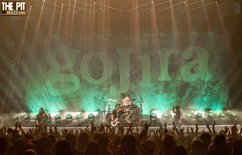 Concert scene showcasing the band Gojira performing on stage with vibrant green lighting and a crowd of fans cheering with raised hands.