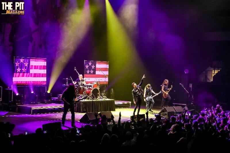 A live rock concert featuring Skid Row performing on stage under colorful lights, with an enthusiastic audience and American flags in the background.