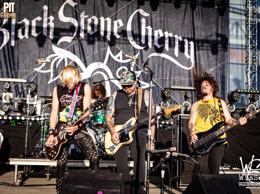 Band members of Black Stone Cherry performing lively on stage with guitars under a large banner with their band name.