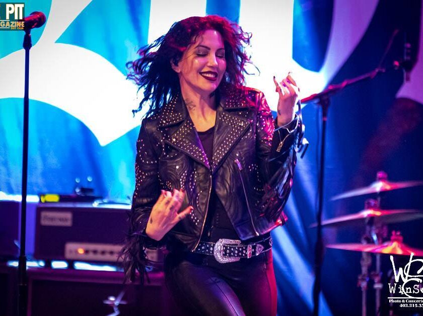 A female singer named Dorothy with curly hair, dressed in a black studded leather jacket, performing energetically on stage with a microphone in her hand.