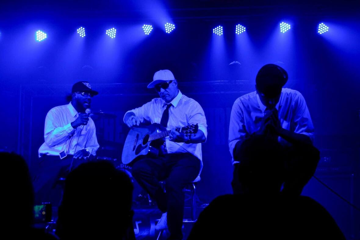 Three men perform on stage, one playing guitar and the other two singing, illuminated by blue stage lights during the "Az The World Burnz" tour.