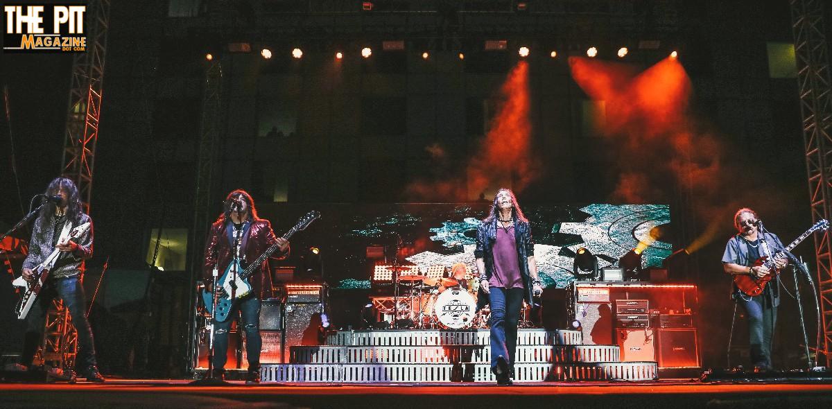 Rock band performing on stage with colorful Tesla lighting and smoke effects, surrounded by music equipment and a large screen in the background.