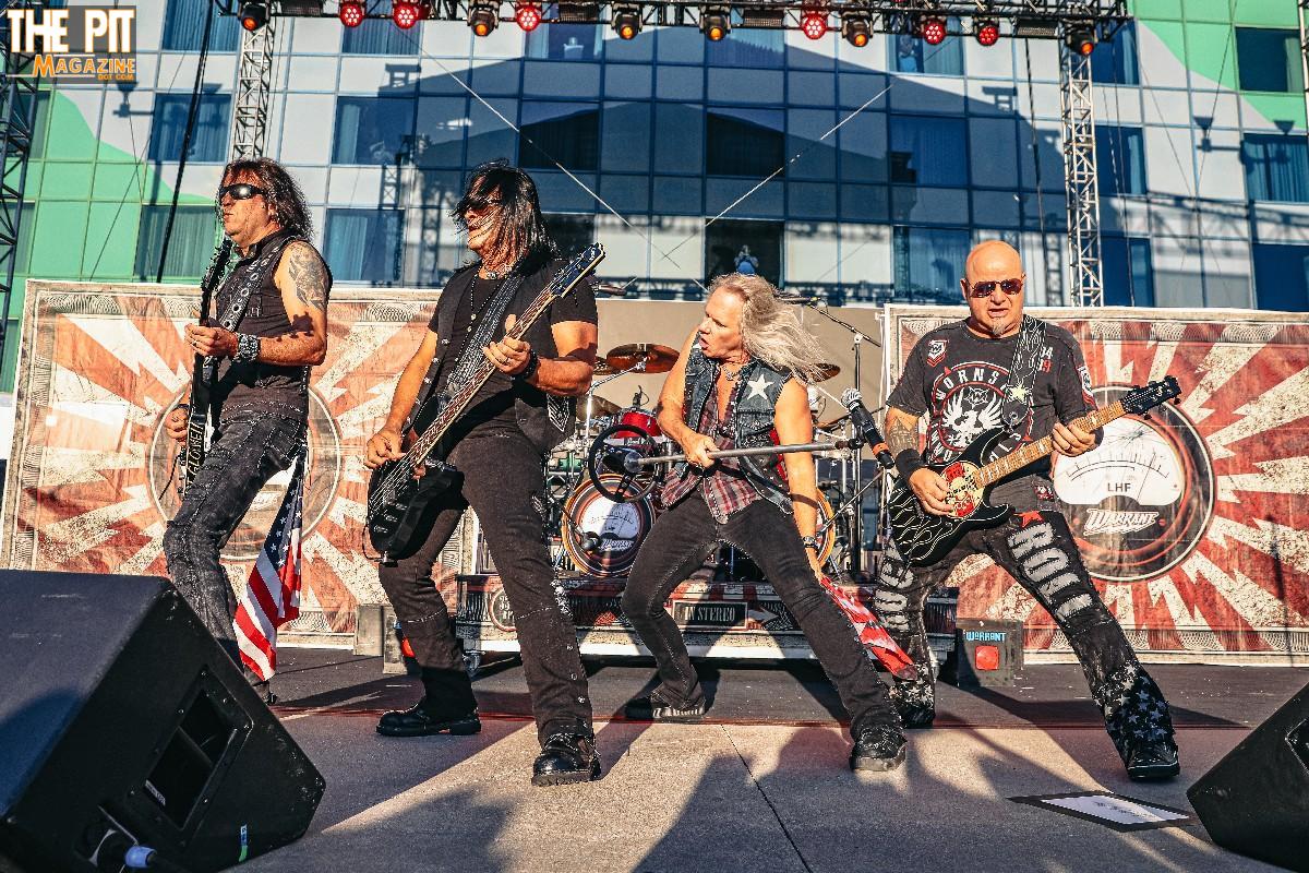 Four rock musicians energetically performing on an outdoor stage, with a large banner displaying Warrant's logo in the background.