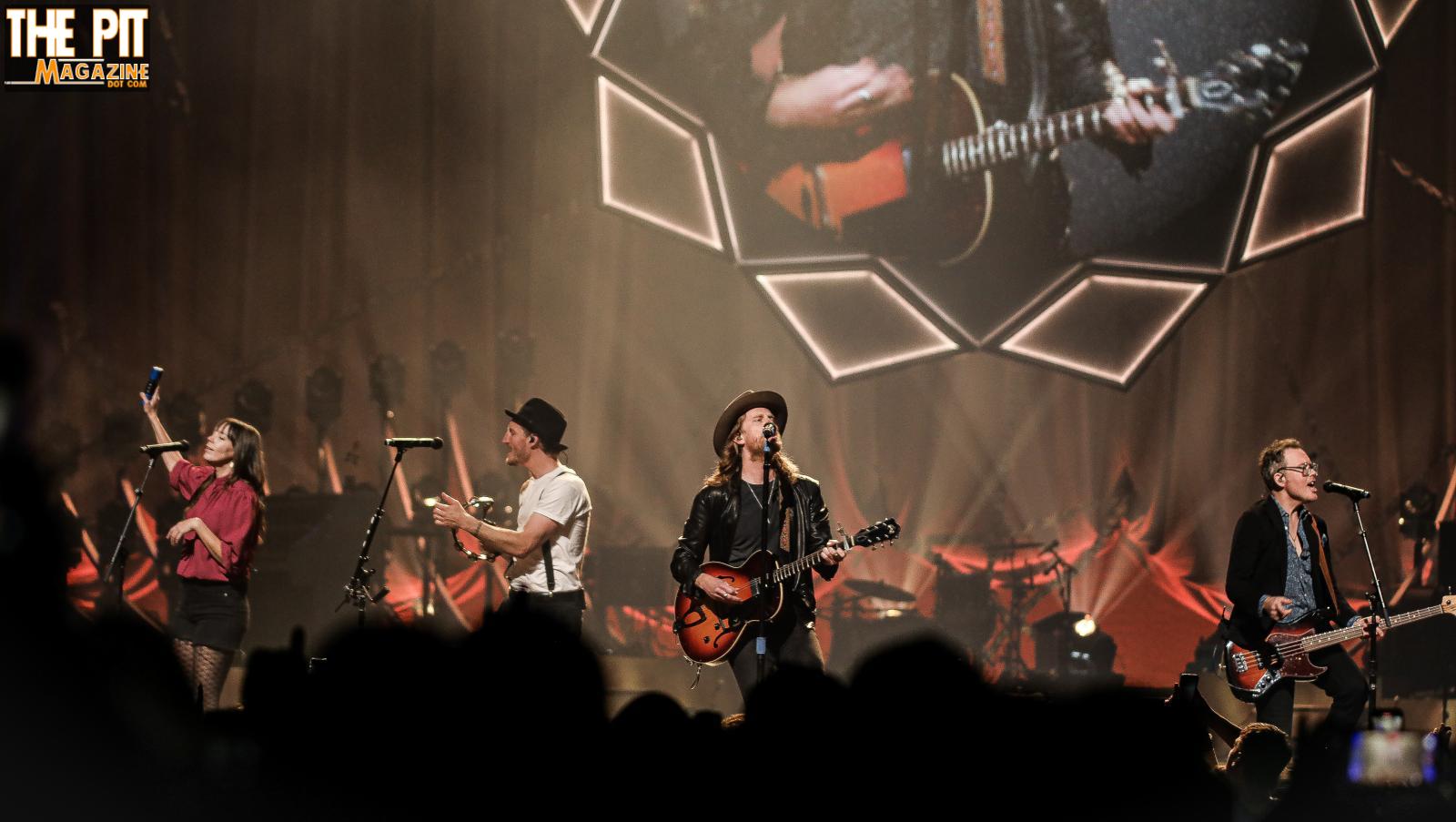 The Lumineers perform on stage, with the lead singer playing guitar and three other members with microphones and instruments, in front of a geometric backdrop.