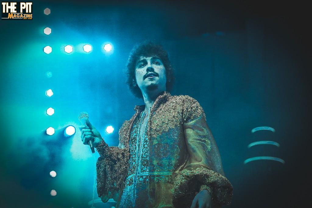 A member of Greta van Fleet in an elaborate golden jacket singing on stage, bathed in blue stage lights with blurred lights in the background.