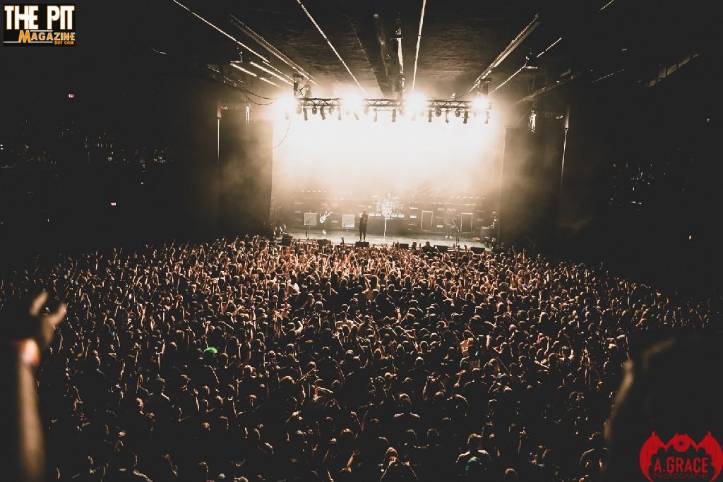 A crowded concert hall with a large audience watching Rise Against perform under bright stage lights.