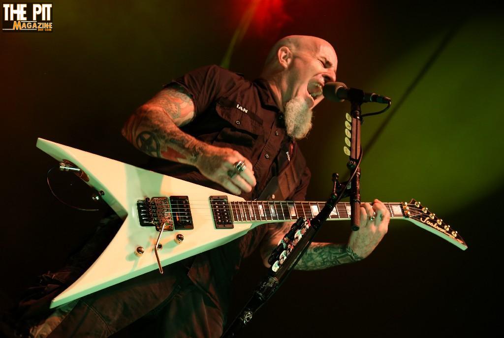 A bald, tattooed male guitarist passionately singing into a microphone while playing a white, angular electric guitar on stage, with green and red stage lighting in the background during an Anthrax concert.